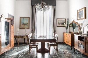 Comedor con piso de mármol, una gran mesa de madera y sillas. Hay una araña colgante sobre la mesa, y la habitación tiene cuadros en las paredes.
