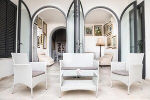 Una habitación con tres sillas blancas de mimbre, una mesa de centro de vidrio y un sofá con un cojín gris.