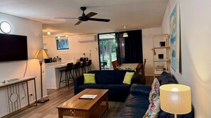 Una sala de estar con sofá, mesa de centro y televisor. Hay una zona de cocina con mesa y sillas.