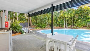 Un espacio al aire libre cubierto con piscina, muebles blancos y abundante vegetación.