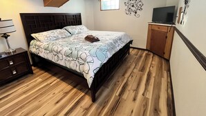 A bedroom with a bed, wooden floor, a nightstand, a lamp, a TV, and a decorative wall piece. - Spacious 3 level 4-bedroom house- 2 Full kitchens-2 king beds and 3 queen beds. (Campbell)