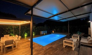 Una piscina esterna con un pontile in legno, lettini prendisole e una zona patio coperta.