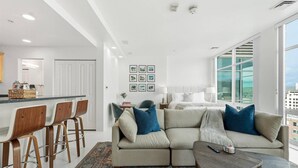 Una sala de estar moderna con un sofá grande, una mesa de centro y una zona de bar con taburetes. Hay cuadros enmarcados en la pared y vistas de la ciudad a través de amplias ventanas.