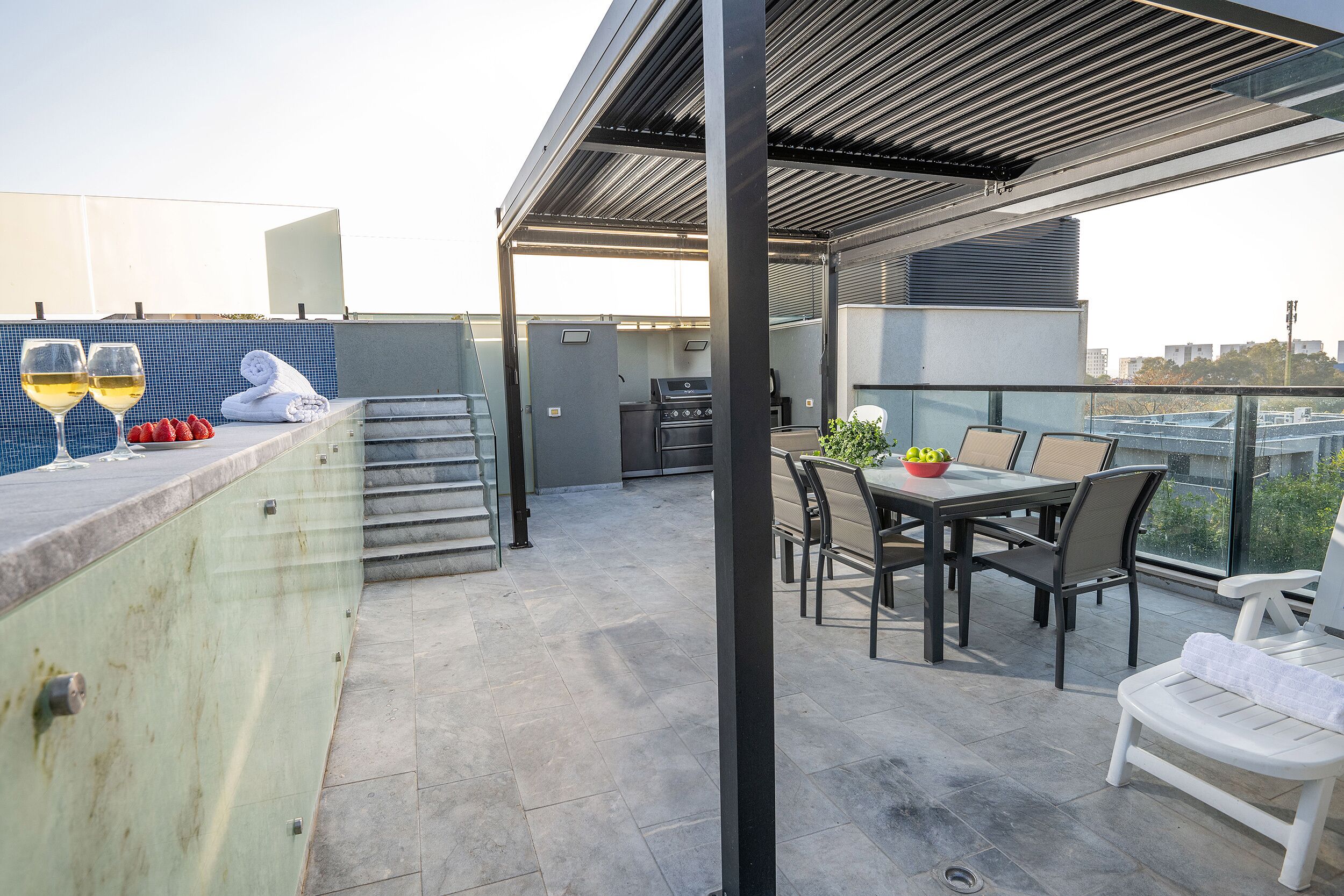Terraza en la azotea con mesa de comedor, sillas, una parrilla y zona de bar con vasos y frutas.