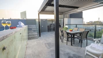 Terraza en la azotea con mesa de comedor, sillas, una parrilla y zona de bar con vasos y frutas.