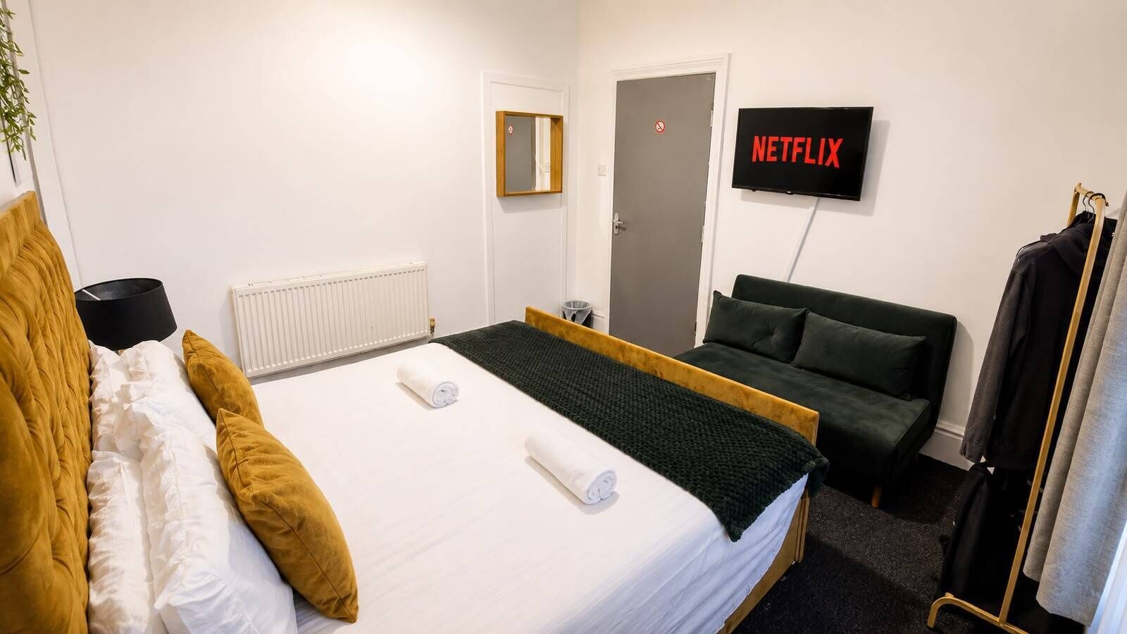 Een hotelkamer met een bed, een groene bank, een televisie waarop 'NETFLIX' wordt getoond, en een kapstok.