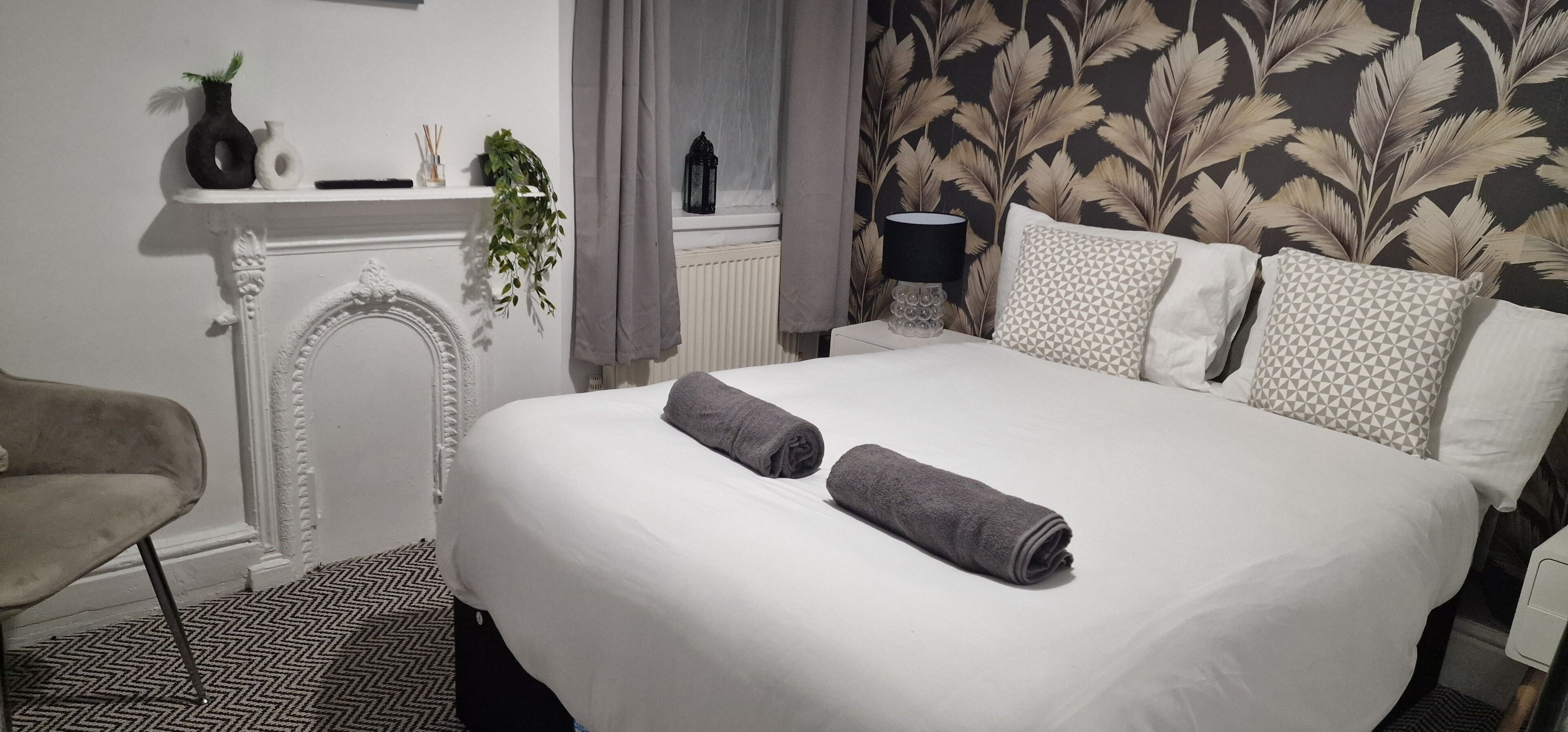 A hotel room with a bed, two towels, a chair, a fireplace, a plant, and patterned wallpaper.