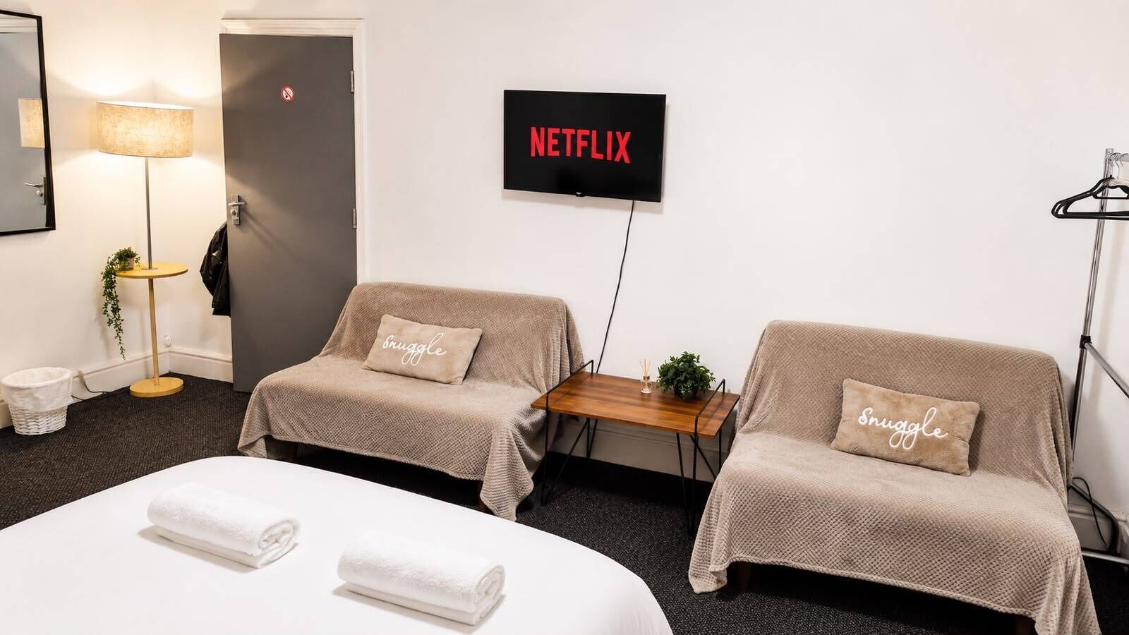 A hotel room with two beds, a TV displaying 'NETFLIX', a small table, and two sofas with 'snuggle' pillows.