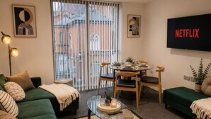 Una sala de estar moderna con un sofá verde, una mesa de centro de vidrio y una zona de comedor con un televisor mostrando 'NETFLIX'.