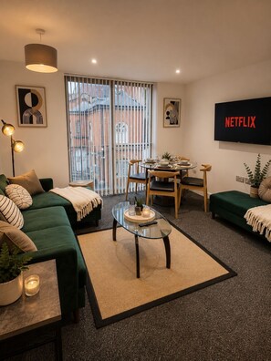 Ein modernes Wohnzimmer mit einem grünen Sofa, einem Glastisch und einem Essbereich, auf dem Fernseher 'NETFLIX' läuft.