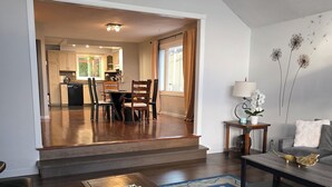 A spacious living room with a wooden coffee table, a blue patterned rug, a leather sofa, and a dining area with wooden chairs and a kitchen in the background. - Lakeview, Warm and Inviting, Family Home (Keswick)