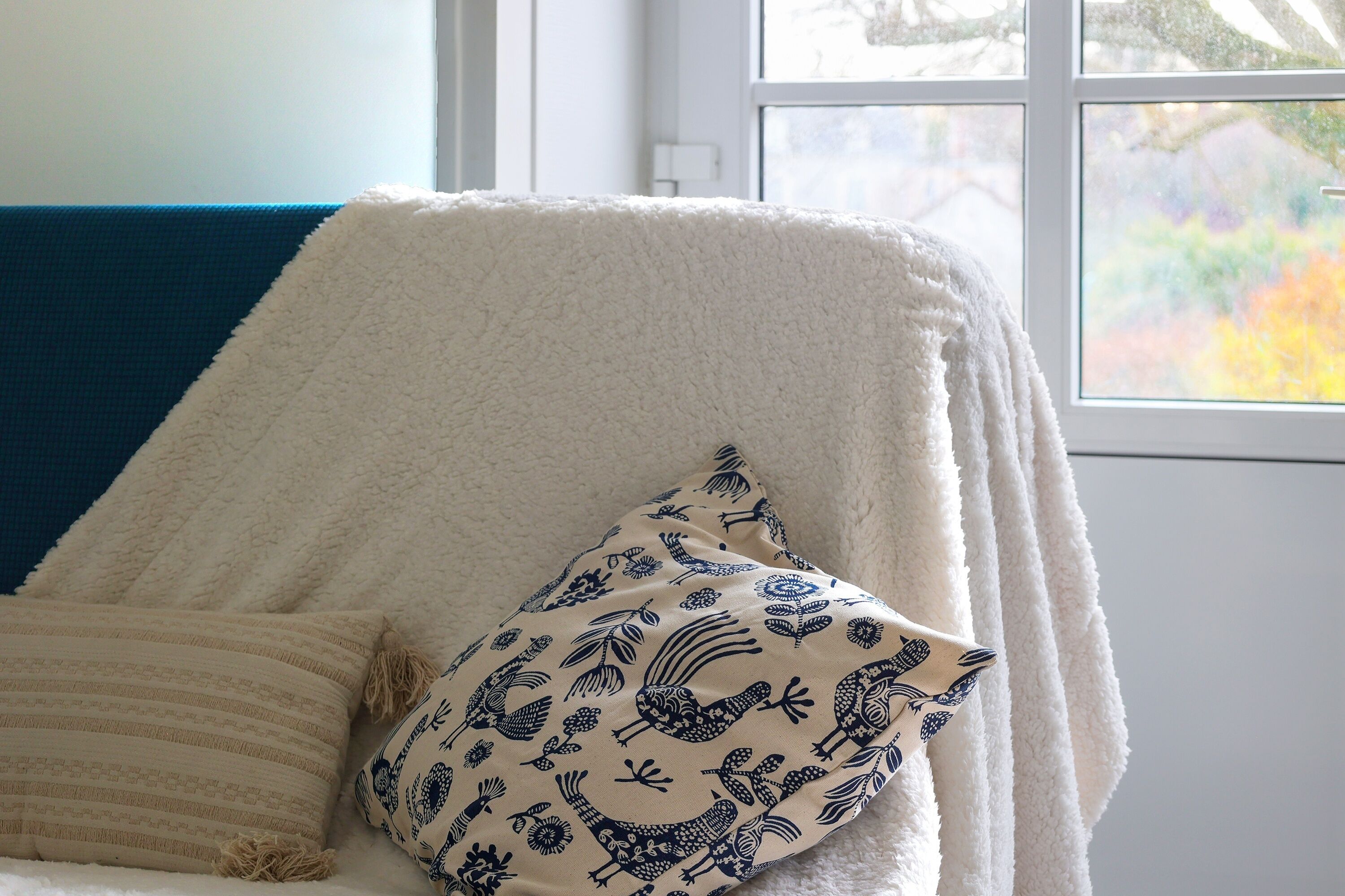Une pièce chaleureuse avec un fauteuil blanc recouvert d’une couverture blanche et moelleuse, un coussin à rayures et un oreiller orné d’un motif d’oiseau.