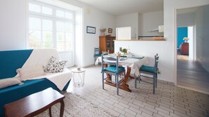 A bright living room with a blue sofa, a dining table with chairs, and a kitchen area visible through an open door. - Le Clos Vendéen - Piscine Privée (L'Hermenault)