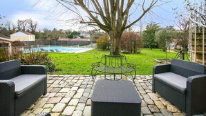 A patio with wicker furniture, a tree, and a swimming pool in the background. - Le Clos Vendéen - Piscine Privée (L'Hermenault)