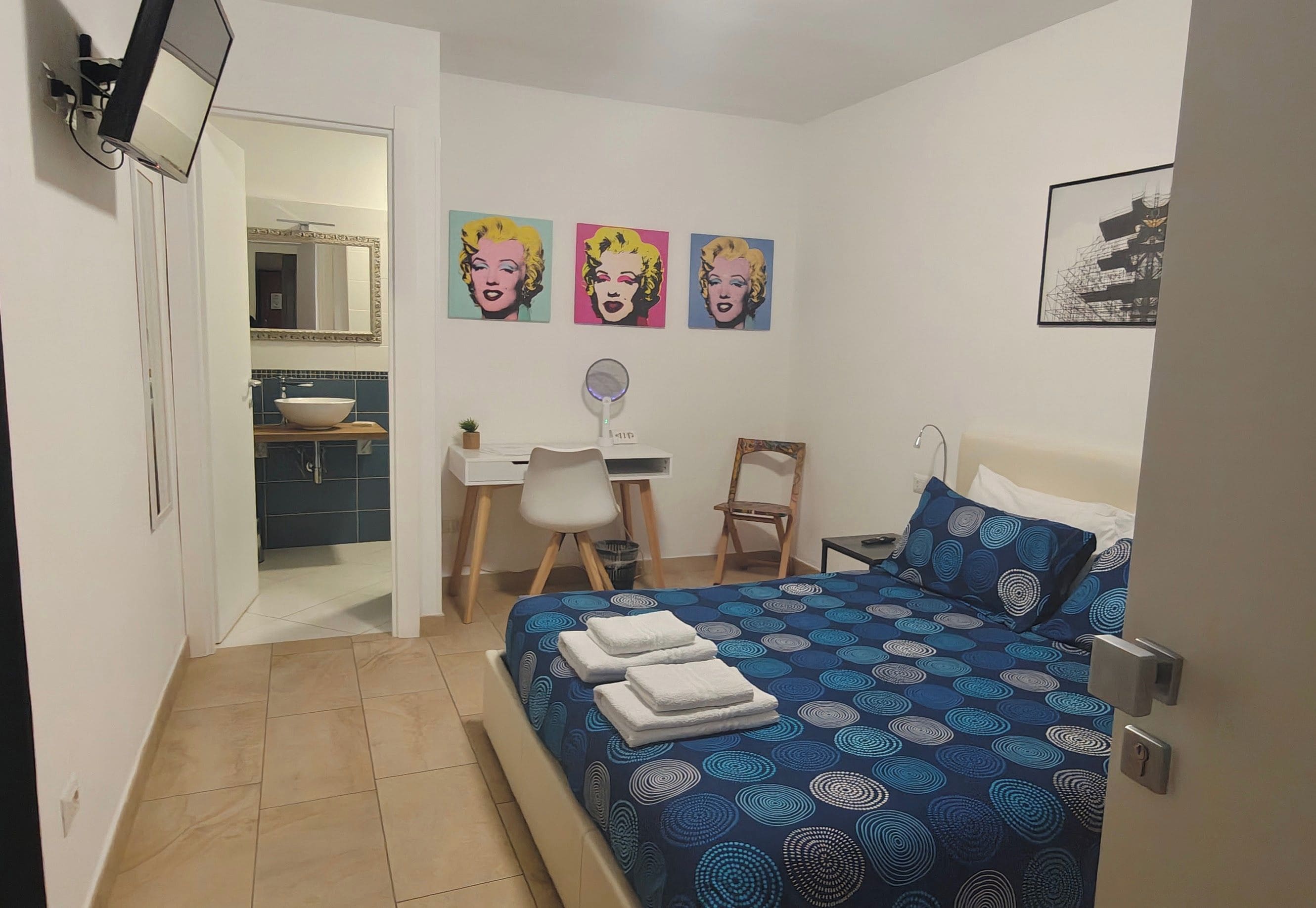 Een slaapkamer met een bed, een bureau, een stoel, een spiegel en kunst aan de muur.