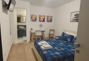 Un dormitorio con cama, escritorio, silla, espejo y obras de arte en la pared.