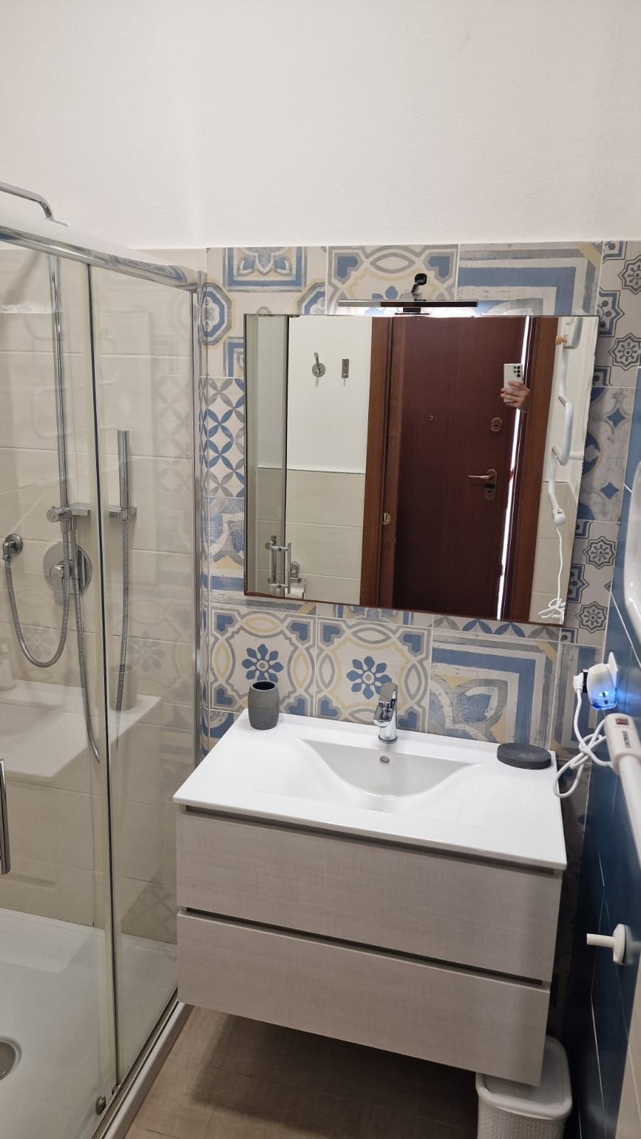 A bathroom with a glass shower enclosure, a white sink with a mirror above it, and patterned tiles on the walls.