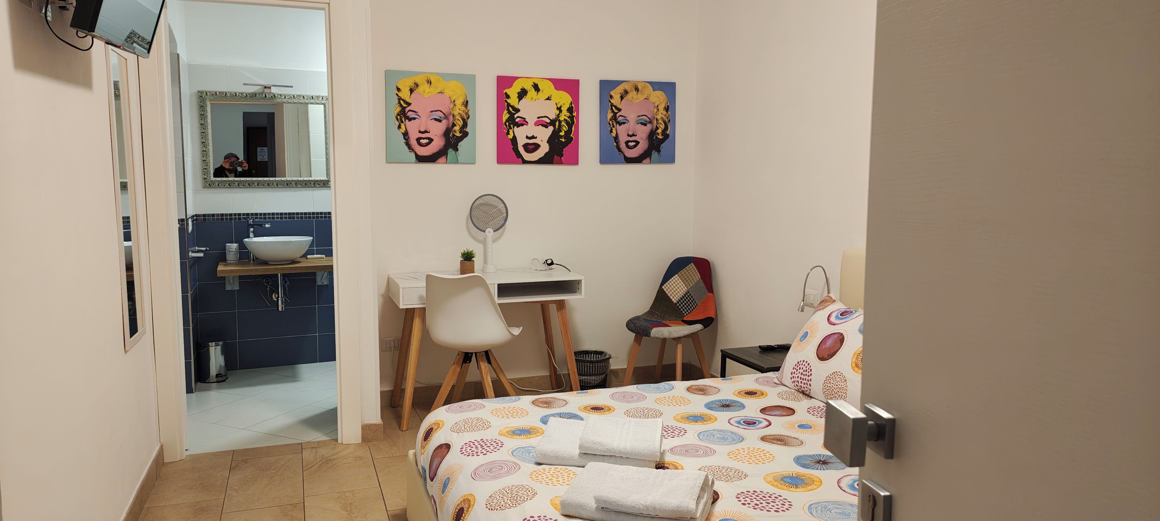 Una camera d'albergo con un letto, una scrivania con una sedia, uno specchio e tre quadri in stile pop art alle pareti.