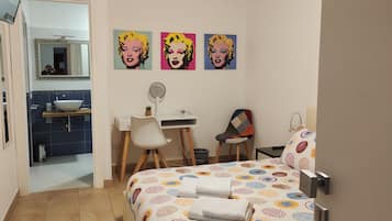 Una camera d'albergo con un letto, una scrivania con una sedia, uno specchio e tre quadri in stile pop art alle pareti.
