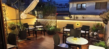 Un patio exterior con varias zonas de estar, incluyendo sillas y mesas, rodeado de plantas en macetas y una valla decorativa.