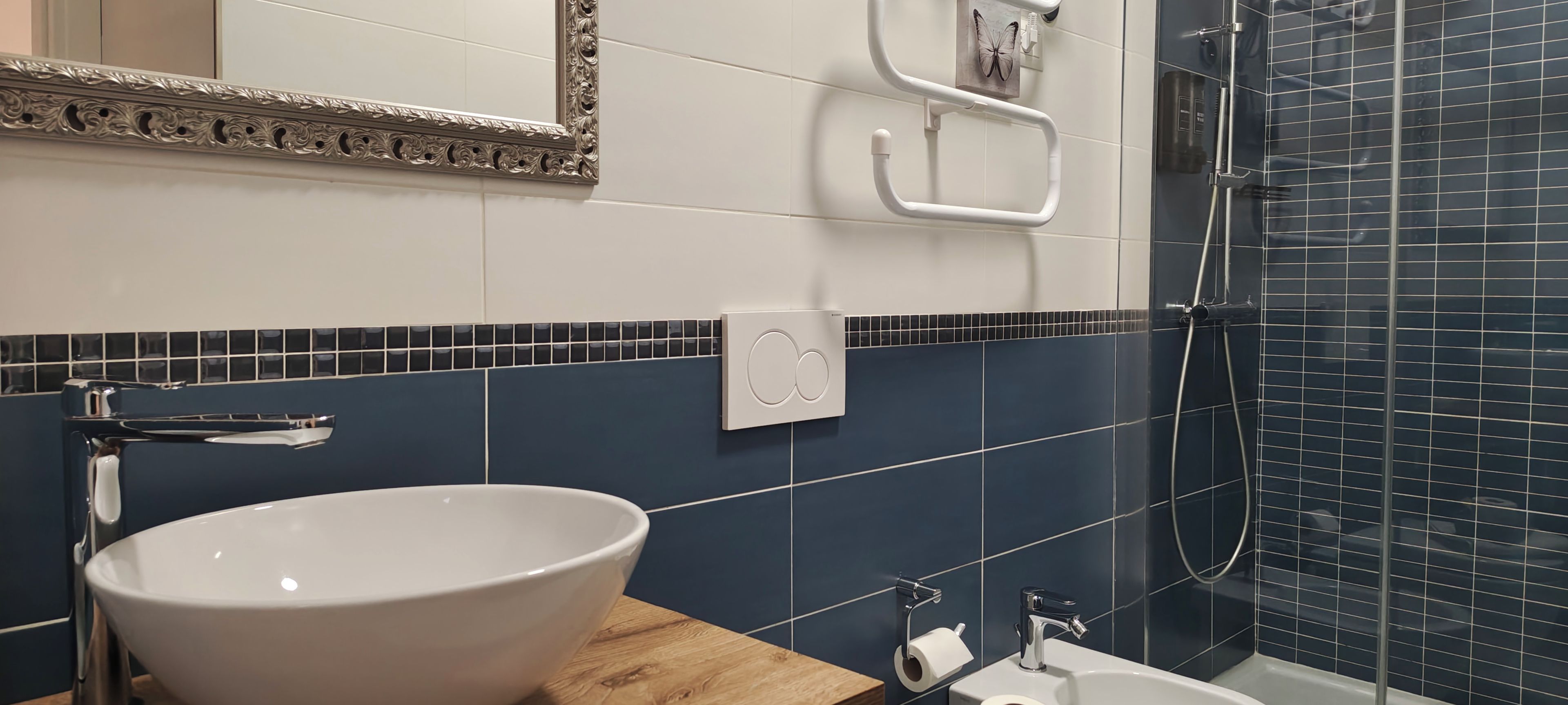 Un baño con un lavamanos blanco, un espejo con marco decorativo, un perchero y una zona de ducha con azulejos azul oscuro.