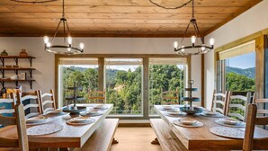 A dining area with wooden tables and chairs, large windows offering a view of the forest, and hanging light fixtures.
