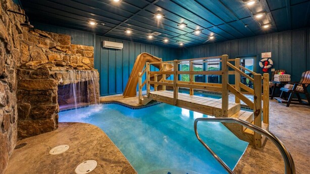 An indoor swimming pool with a wooden bridge, stone wall, and a waterfall feature.