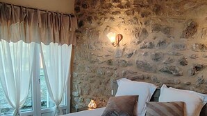 A bedroom with a large bed, stone wall, wooden beams, and a window with curtains. - Gite for 15p at the foot of Mont Ventoux_Lacets du Ventoux (Beaumont-du-Ventoux)