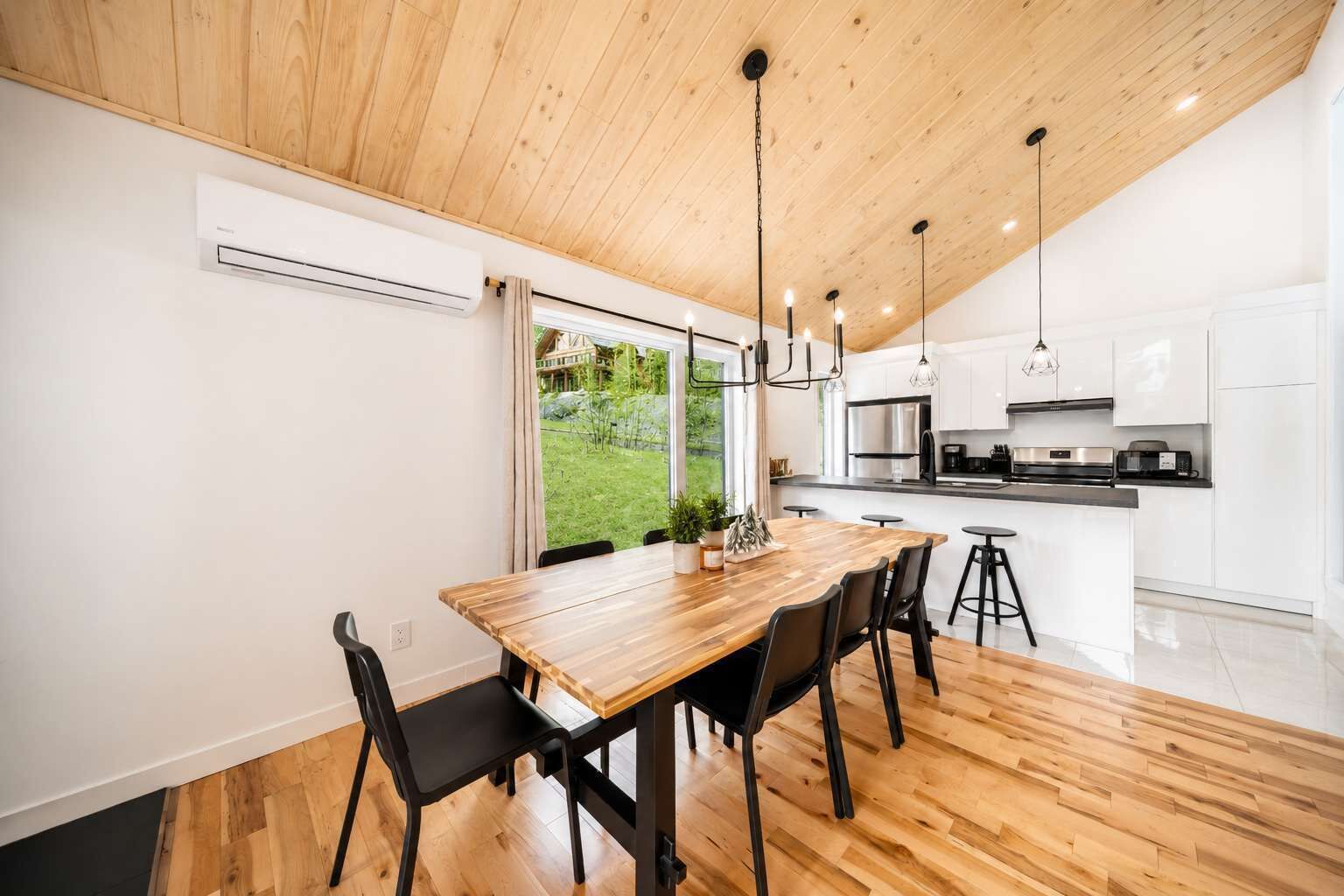 A modern kitchen with white cabinets, a wooden dining table with chairs, and a view of a green garden.