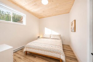 A bedroom with a wooden ceiling, a bed with patterned bedding, a nightstand, a window with a view of trees, and a framed picture on the wall. - Spa & Foosball | Le WKD (Saint-Philémon)