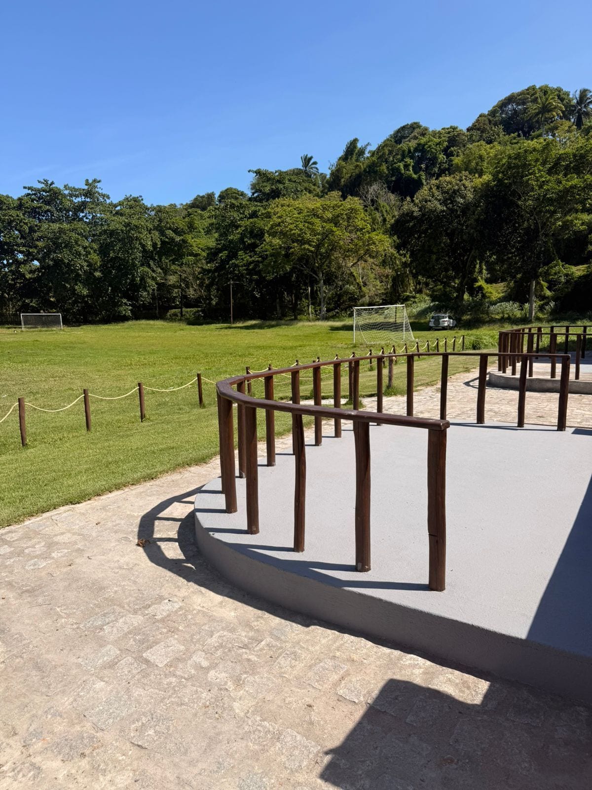 Un chemin pavé, bordé de poteaux en bois et délimité par une corde, mène à une zone herbeuse où se trouve un but de football.