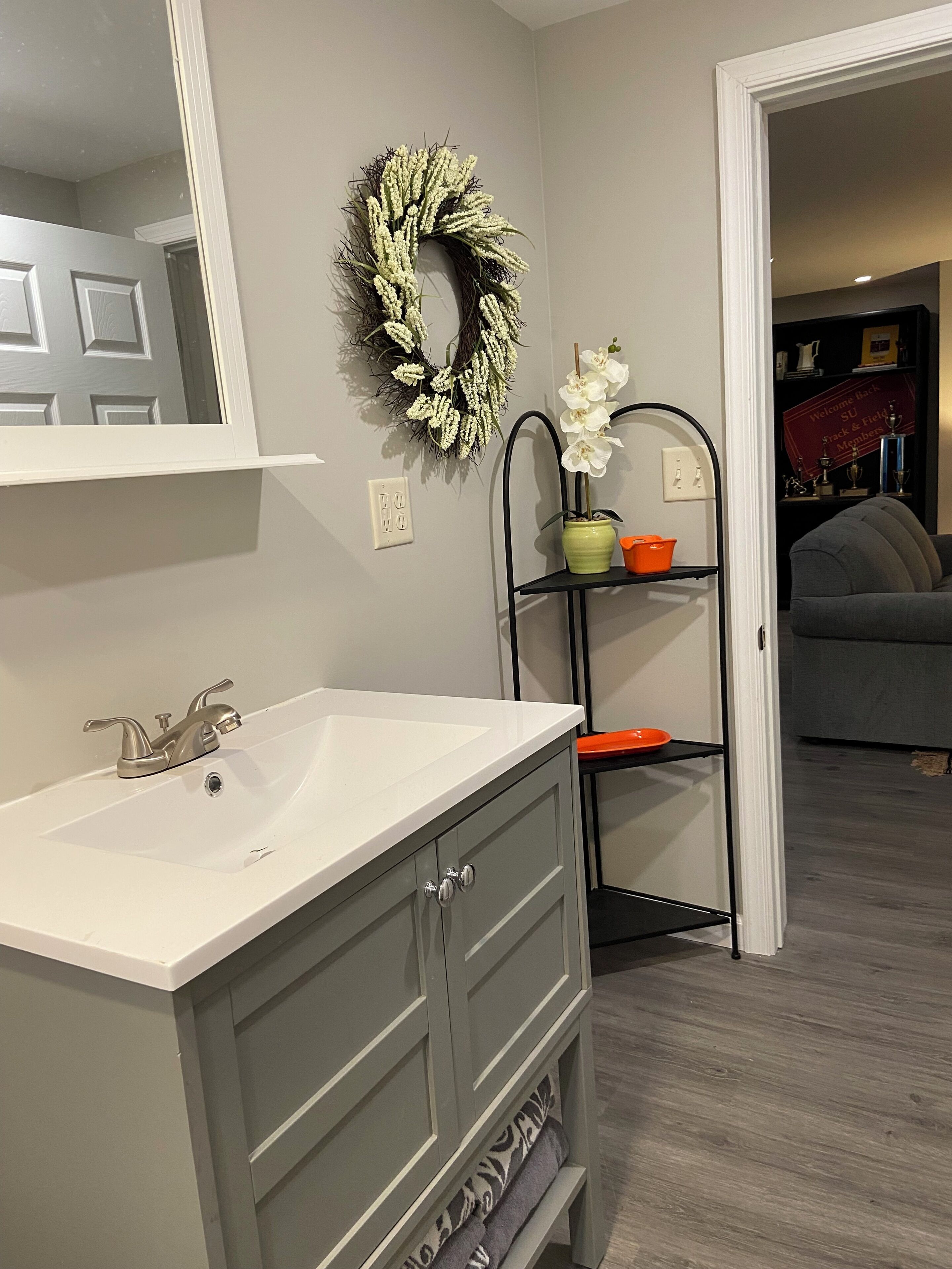 A bathroom with a white sink, a mirror, a gray vanity, a black shelf with a potted orchid, and a wreath on the wall.