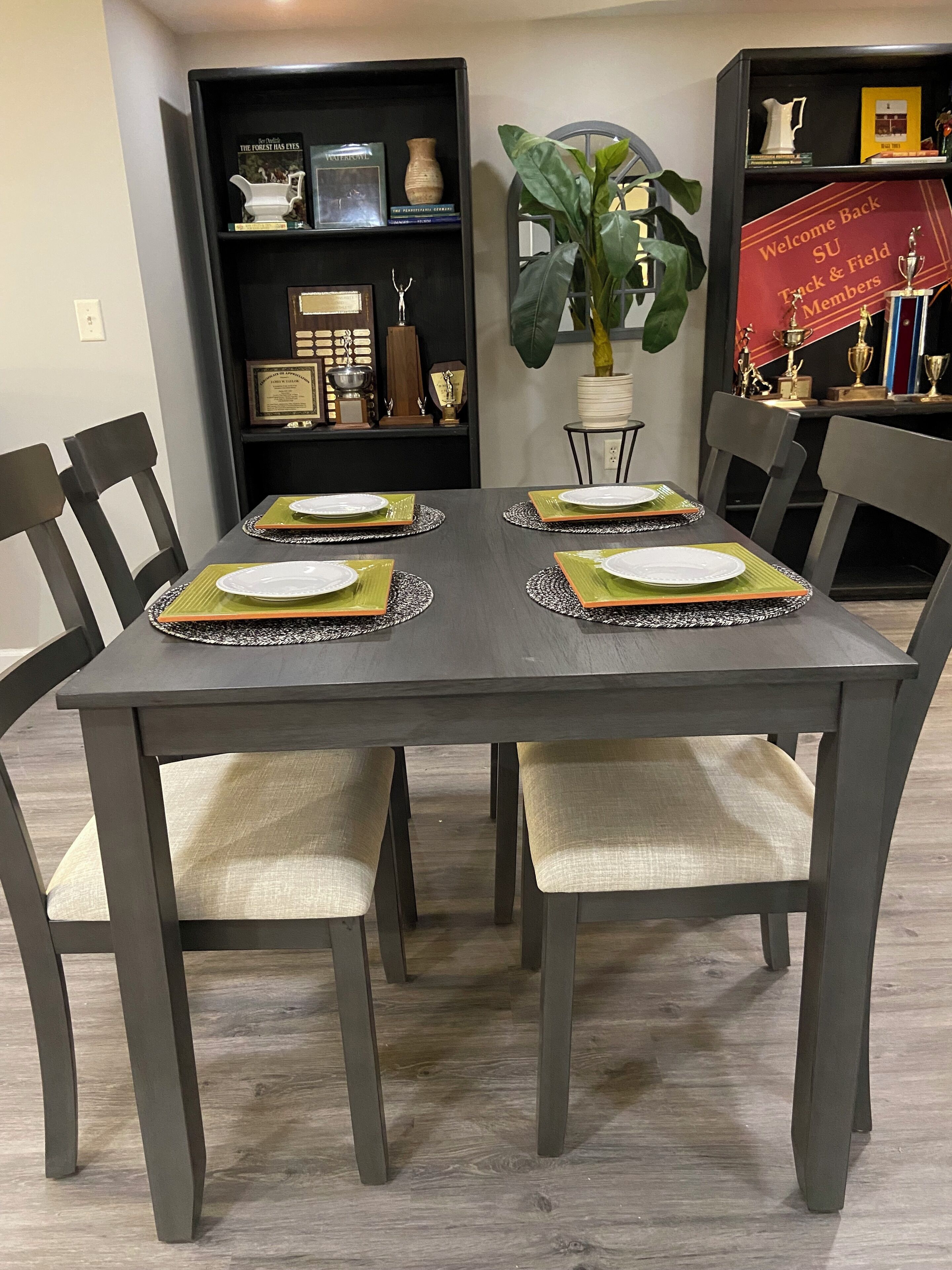Een eettafel gedekt met vier borden, elk met een groene placemat. Rond de tafel staan zes stoelen, en er is een boekenkast met verschillende spullen op de achtergrond te zien.