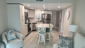 A modern kitchen with white cabinets, a stainless steel refrigerator, and a dining area with a white table and chairs. - IT'S A SHORE THING - 
ON THE BEACH - SPECTACULAR SUNSETS
NEWLY RENOVATED (Fort Myers Beach)