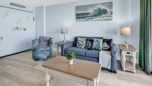 A living room with a sofa, two side tables with lamps, a coffee table with a plant, and a wall with a wave painting and seagulls. - Compass Cove 366 (Myrtle Beach)