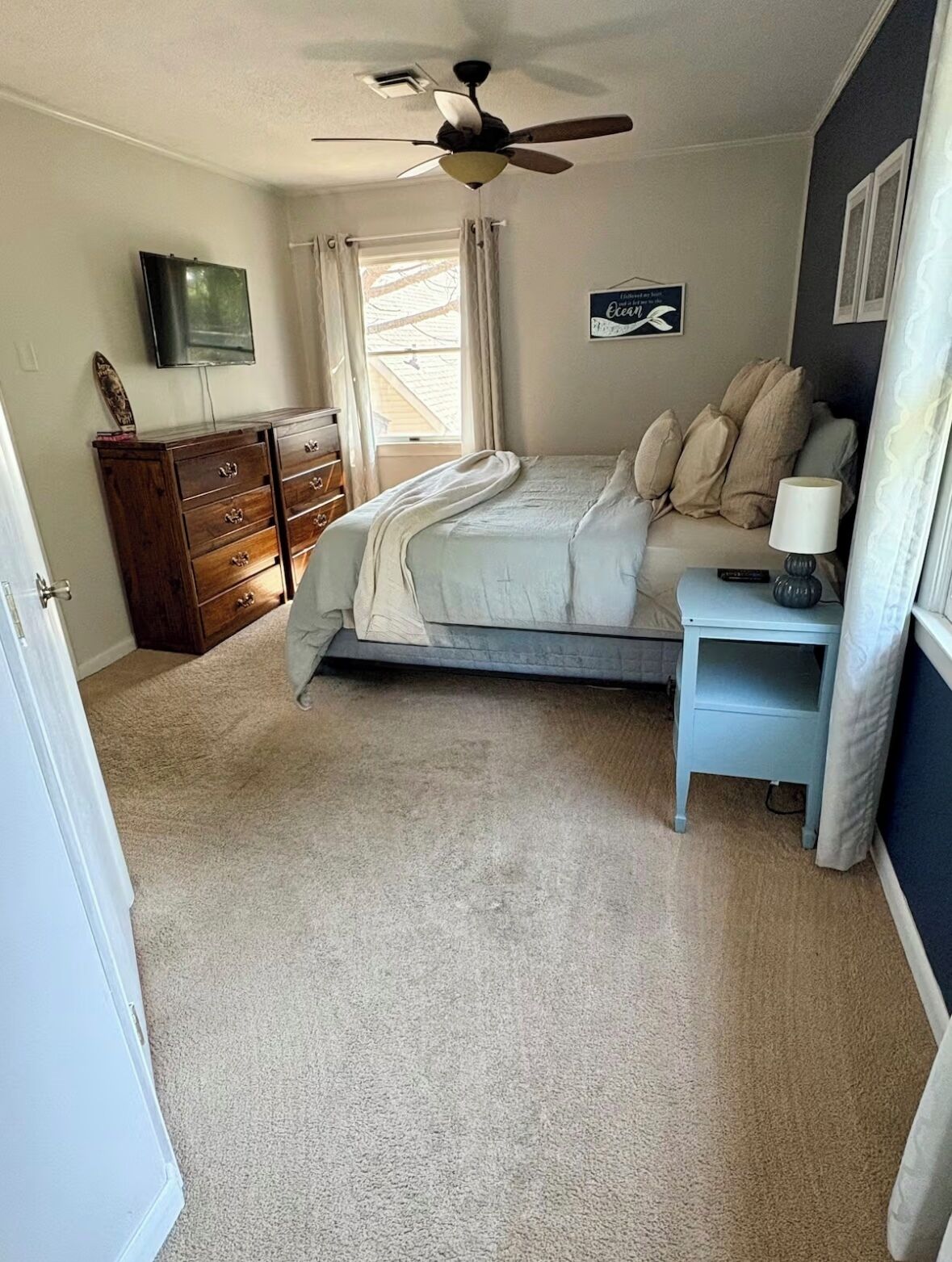 A bedroom with a bed, a nightstand, a dresser, a ceiling fan, a TV, and a window with curtains.