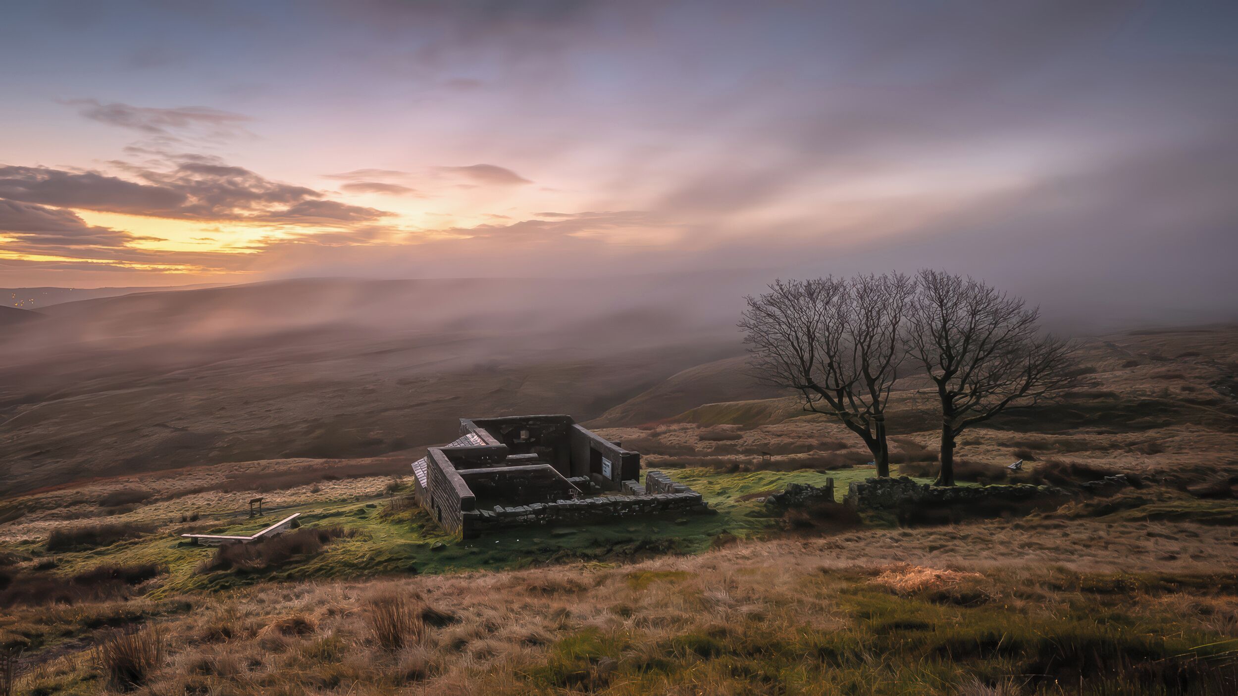 A stone structure with a bench, surrounded by grass and a tree, overlooking a valley.