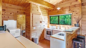 A compact kitchen with wooden walls, a window with a view of greenery, and basic amenities like a refrigerator, stove, and sink. - Kereru View - Lake Taupo Holiday Home (Pukawa)