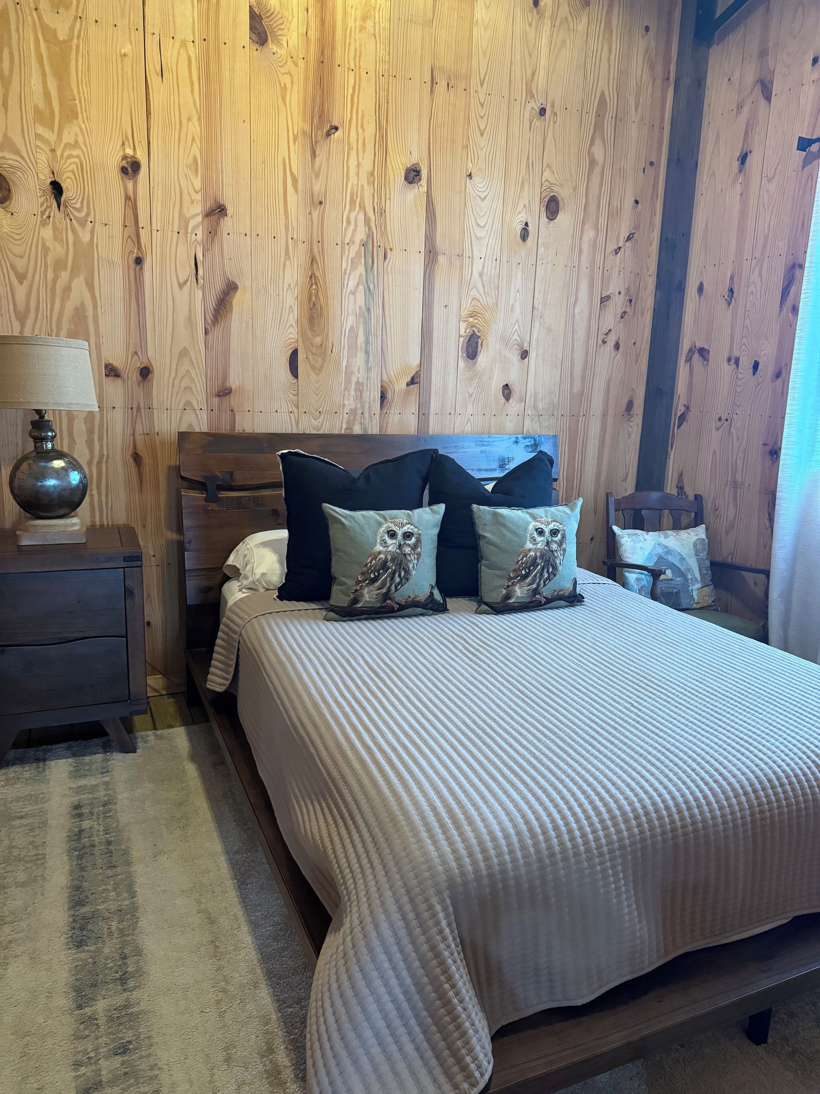 A bedroom with a wooden wall, a bed with a white quilt, and three pillows with owl designs.