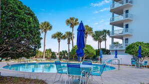 A pool area with blue chairs and umbrellas, surrounded by palm trees and a white building with balconies. - Harbor Landing 102b (Destin)