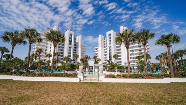 A modern hotel with multiple white buildings, palm trees, and a grassy area with a pool. - Harbor Landing 102b (Destin)