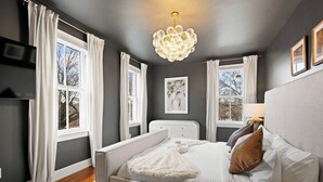 Ein Schlafzimmer mit einem Bett, weißen Vorhängen, einer Kristallleuchter und einem gerahmten Bild an der Wand.