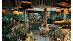Un área de estar al aire libre con mobiliario de mimbre, una jardinera central y luces colgantes.