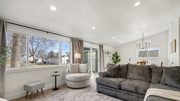 A spacious living room with a large window, a grey sofa, a round ottoman, and a patterned rug.