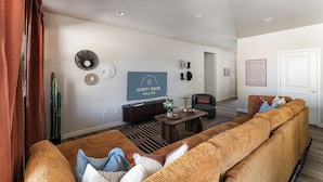A living room with a large sofa, a coffee table, and a flat-screen TV. There are wall decorations and a cactus on the wall. - Dusty Sage Hollow ~ Hot Tub + Arcade + Fire Pit (Fernley)