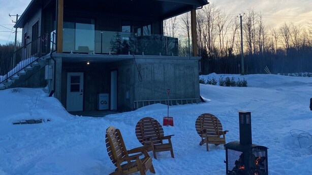 A modern house with a large deck, wooden chairs, and a wood-burning stove in a snowy setting. - Chalet L'Ancrage; anchoring yourself in nature! (Grandes-Piles)