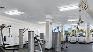 Ett gym med olika träningsmaskiner, ett brandlarm som är monterat i taket och utsikt över palmer och byggnader utanför.