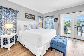 A bedroom with a bed, a nightstand, a blue bench, a window with curtains, and a view of the outside. - Nana's Beach House (Destin)