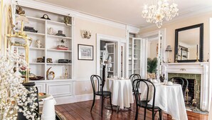 Un comedor con piso de madera, paredes blancas y chimenea. Hay tres mesas de comedor con sillas, un candelabro y estantes con varios objetos.