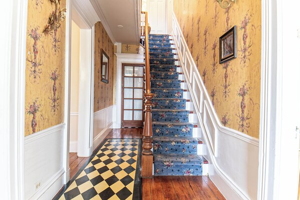 A staircase with floral carpet and a checkered floor mat. - Miss Murphy's High Street Inn (Galena)
